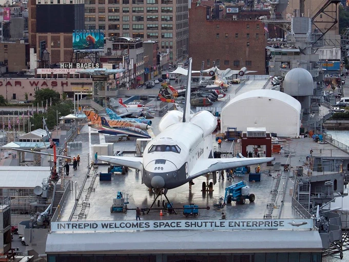 The image shows the space shuttle Enterprise displayed on the deck of the USS Intrepid, a museum ship. Several aircraft are also visible on the deck. Buildings of New York City can be seen in the background. A sign reads "Intrepid Welcomes Space Shuttle Enterprise."