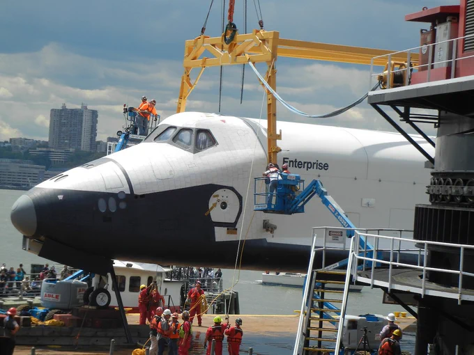 The image shows the space shuttle Enterprise being lifted by a crane near a waterfront. Workers in safety gear are on the platform and lifts, guiding the process. A cityscape and waterfront are visible in the background.