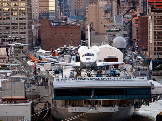 The image shows the space huttle Enterprise displayed on the deck of the USS Intrepid aircraft carrier, part of the Intrepid Museum in New York City. The ship is docked at a pier with the skyline of the city in the background. The banner reads "Intrepid Welcomes Space Shuttle Enterprise." Other aircraft are visible on the deck.
