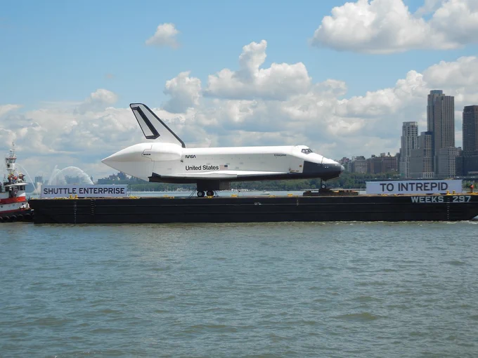 A space shuttle labeled "United States" and "NASA" is displayed on a barge in a body of water. The barge is named "Weeks 297" and carries a banner reading "Shuttle Enterprise to Intrepid." City skyscrapers and a small tugboat are visible in the background.