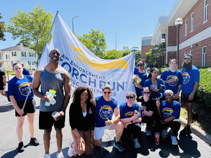 Group of runners posing outdoors in matching blue LETR shirts, holding a large Law Enforcement Torch Run banner on a sunny day.