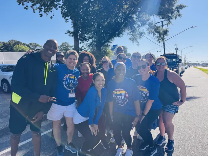 Group of runners wearing matching blue LETR shirts posing together outdoors on a sunny day near a street and trees.