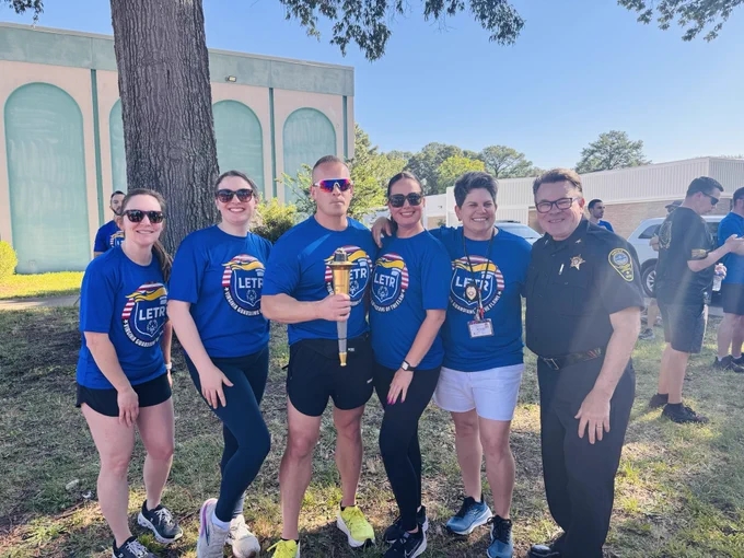 Group of six people standing outdoors on grass, five wearing matching blue LETR shirts and one in a police uniform, holding a torch under a tree.