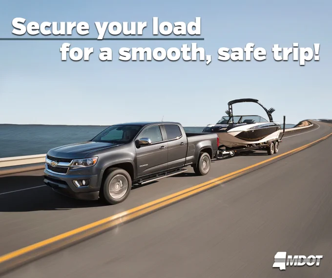 A pickup truck is towing a boat along a road beside a body of water. The image includes the text "Secure your load for a smooth, safe trip!" and an MDOT logo in the bottom right corner.