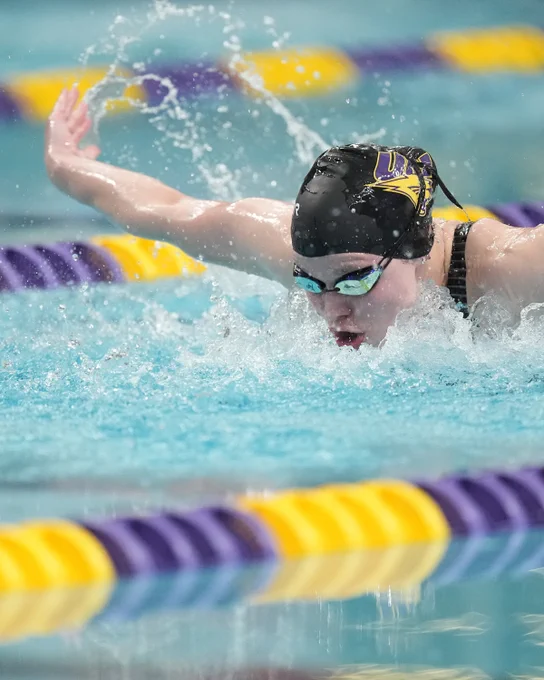 A swimmer in a black cap and goggles competes in a pool, creating splashes. Purple and yellow lane dividers are visible.