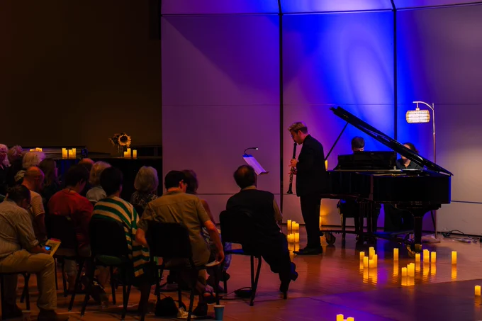 A person plays the clarinet on a dimly lit stage beside a grand piano. The audience sits in rows, surrounded by decorative candles and soft blue lighting.