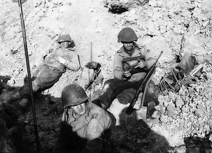Three soldiers in helmets are positioned in a trench, with one reading a book and the others looking vigilant, in a stark black and white wartime photograph.