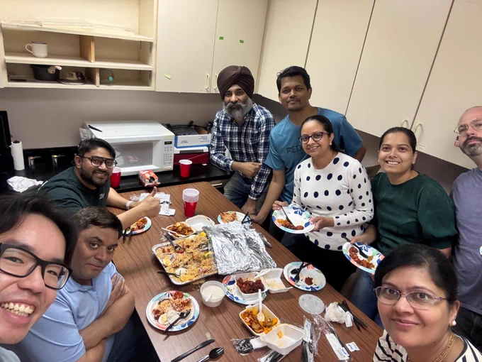 A group of people are gathered around a table in a kitchen setting, enjoying a meal together. There are takeout containers and plates filled with food. The individuals are smiling at the camera, and a microwave and cabinets are visible in the background.