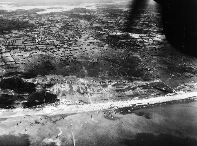 Aerial black and white photograph of the D-Day landings at Normandy, showing the coastline and patchwork fields with visible military activity.