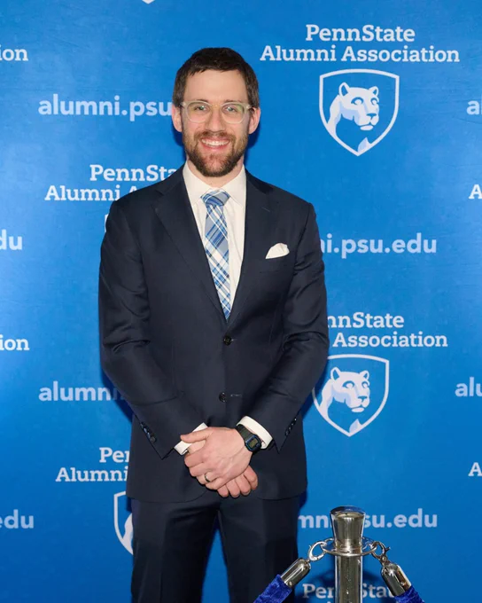 Matthew Kapelewski posing in front of a backdrop with the Penn State Alumni Association mark on the back at the Alumni Achievement Award ceremony.