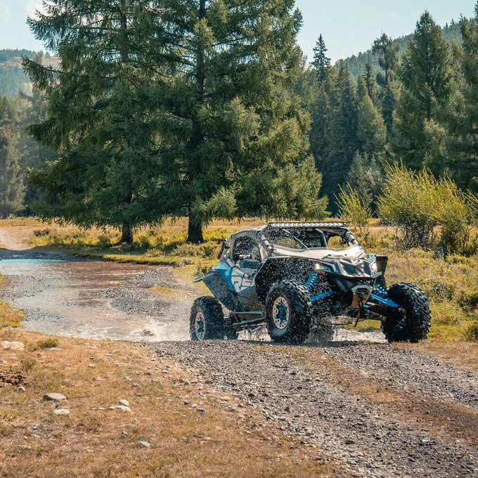 An off-road vehicle drives on a rugged path through a forested area with tall trees, slightly splashing water from a shallow stream. The setting is natural and serene.