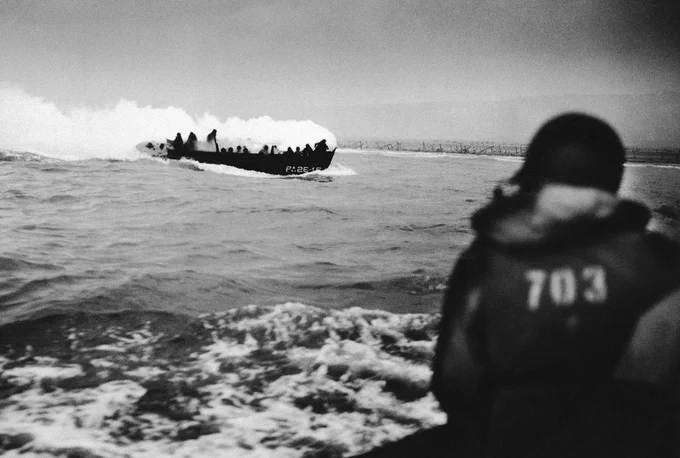 A historical black and white photo showing a lifeboat in choppy seas, filled with people and emitting smoke, as another person in a life vest labeled "703" watches on from the foreground.