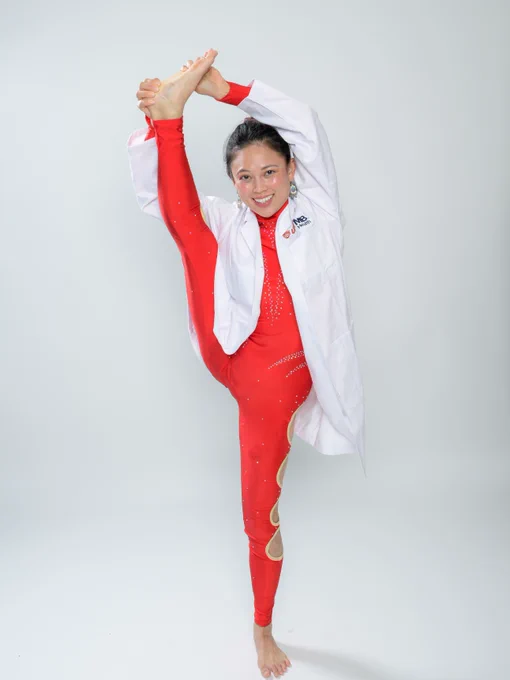 A person wearing a red gymnastic outfit and a white coat with the UTMB Health logo performs an impressive vertical leg stretch, smiling against a plain background.