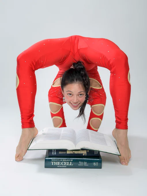 A person in red, flexible attire is performing a contortion pose, balancing on two open science books, titled "Microbiology" and "The Cell." The person is smiling and appears to be in a studio setting.