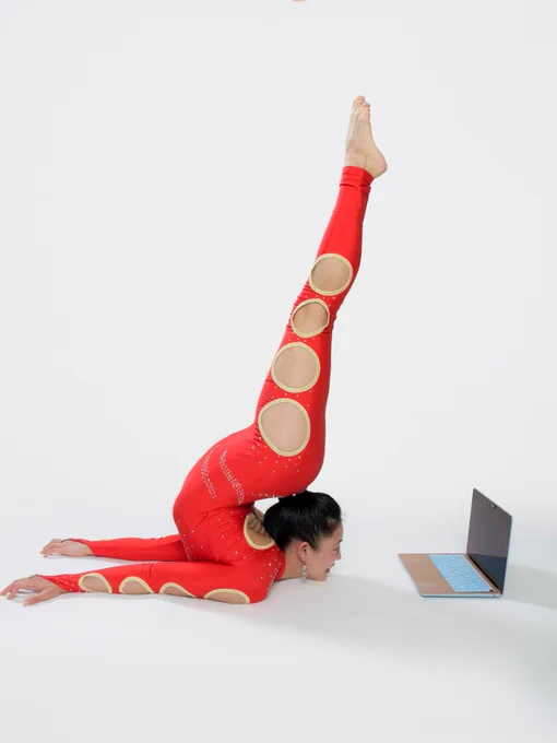 A person in a red, circular cut-out bodysuit performs a yoga pose with legs extended vertically while using a laptop on the floor.