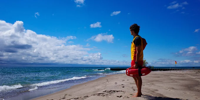 Lifeguard in yellow and red gear standing on a sandy beach, watching the ocean under a partly cloudy sky.