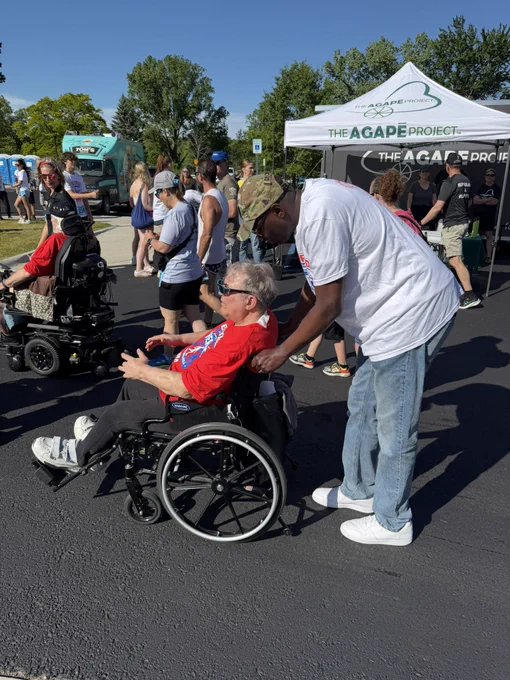 Lieutenant Governor Garlin Gilchrist II, wearing a camouflage cap and white t-shirt, leans forward on an outdoor asphalt lot to push a man wearing sunglasses and a red shirt in a wheelchair. Other event participants and a white tent branded "The Agape Project" are visible in the background.