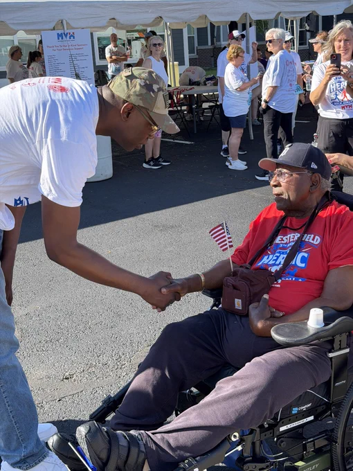 Close-up profile view of Lieutenant Governor Garlin Gilchrist II leaning over to shake hands with an elderly Black man wearing a Detroit Tigers baseball cap and sitting in a wheelchair. The man in the wheelchair is wearing a red event shirt and holding a small American flag. Other attendees stand under a white event tent in the background.