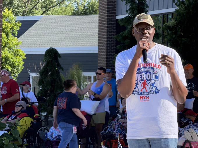 Michigan Lieutenant Governor Garlin Gilchrist II speaking into a microphone outdoors. He is wearing glasses, a camouflage baseball cap, and a white t-shirt that reads "Chesterfield 5th Annual Heroic Walk, Run & Roll." In the blurred background, several community members are gathered, including individuals using wheelchairs.