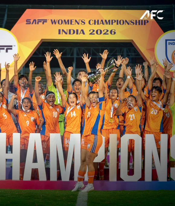 The Indian women's football team celebrates their victory at the SAFF Women's Championship 2026. The players are jubilant, holding up the trophy and cheering under the championship banner.