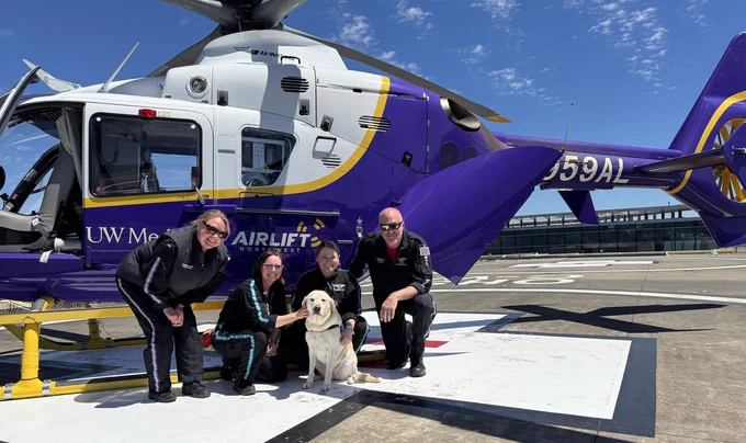 A group of people and a dog are on a helipad next to an Airlift Northwest helicopter. The helicopter is branded with "UW Medicine." The sky is clear and sunny.