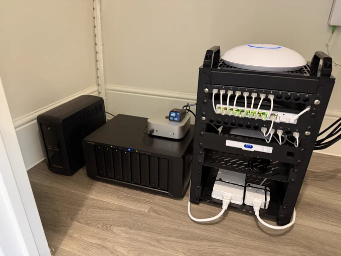 A photo of a homelab setup showing a, from left to right, UPS, NAS with a Mac mini sitting on top of it and finally a black mini rack with a bunch of networking equipment mounted in it