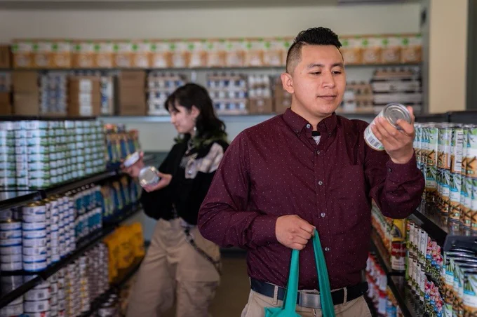 Two California community college students stand side by side inside a campus basic needs store, thoughtfully exploring a selection of neatly arranged canned food items on fully stocked shelves. The space is clean and organized, with rows of brightly labeled cans creating a sense of accessibility and abundance. Their focused expressions reflect both curiosity and reassurance, highlighting the supportive resources available to help students meet their basic needs while staying on track with their education.
