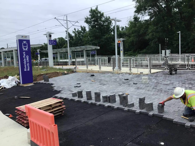 Construction worker laying gray paving stones at a transit station platform under cloudy sky.