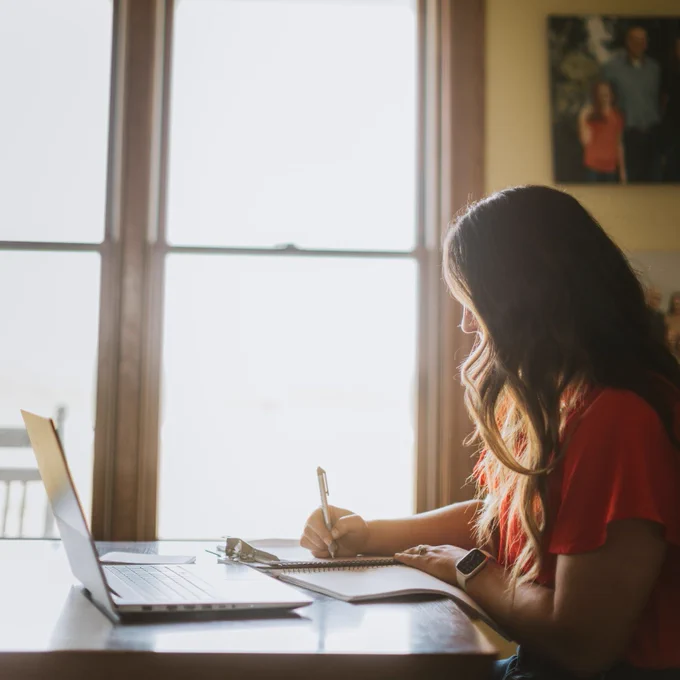 Female farmer sits at table working with computer open and taking notes in a notebook. 