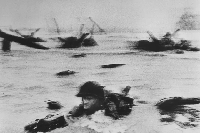 Soldiers wading through water with military equipment in the background during the D-Day Normandy landings.