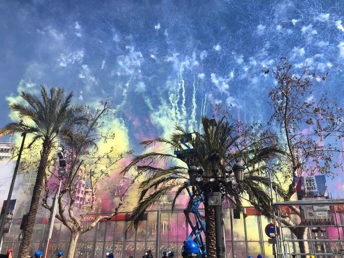 Plaza urbana con palmeras y humo multicolor de fuegos artificiales, andamios y personas con cascos azules.