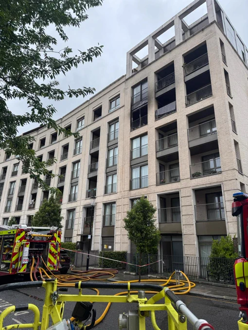 A block of flats with some evidence of fire damage on the building. In front of the building is the rear of a fire engine with hoses on the ground.
