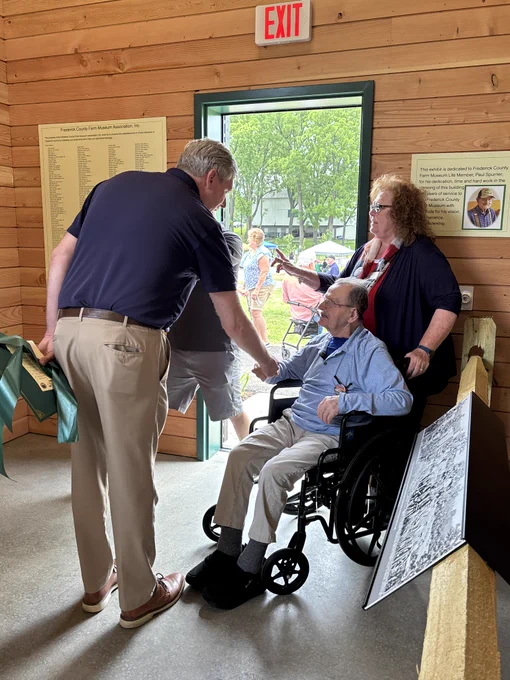 Inside a wooden‑walled barn, a person seated in a wheelchair is positioned near the entrance where natural light streams in through an open doorway. Two individuals stand nearby, leaning in and engaging with the person in the wheelchair. The person in the wheelchair is wearing light-colored pants, dark socks, and darker footwear, along with a long-sleeve shirt. The standing individuals wear casual clothing, including a short-sleeve collared shirt and khaki pants. Behind them, a large display board featuring historical black‑and‑white photos is propped up against a wooden railing. On the wall to the right is a mounted plaque containing several paragraphs of text, and next to it is an EXIT sign above a doorway. Through the open entrance, more people and green trees are visible outside in a shaded area. 
