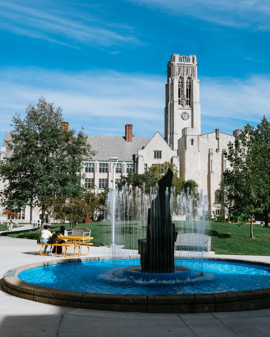 University Hall, set against a clear blue sky. In the foreground, a circular fountain with cascading water is surrounded by a paved walkway. Nearby, two people sit at a yellow picnic table under tree shade. Lush greenery complements the scene.