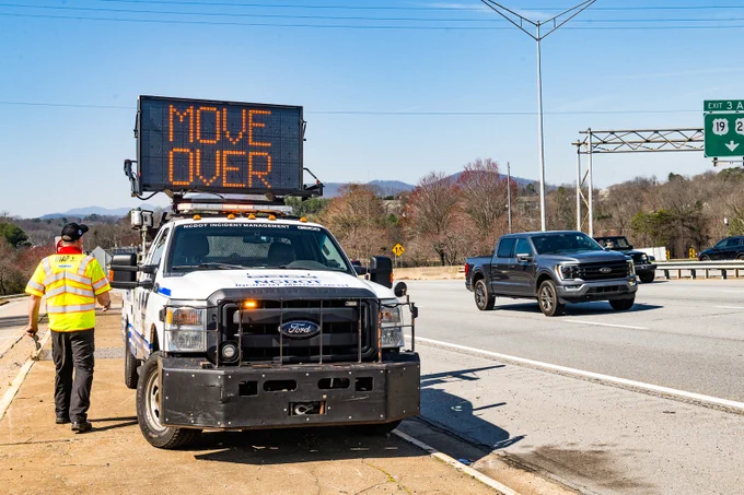 A roadside scene shows a vehicle displaying an electronic sign that reads "MOVE OVER." This vehicle is parked on the shoulder of a highway near Exit 3 for Highway 19. A person stands near the vehicle, which is a Ford truck. Traffic flows on the highway, with trees and distant hills in the background.