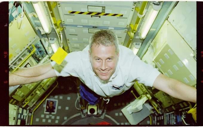 Jay Buckey grins as he flies through a module aboard the space shuttle with arms outstretched.