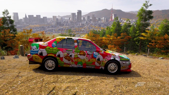Un coche decorado con diseños coloridos de personajes de dibujos animados está estacionado en un camino de tierra. En el fondo, hay un extenso paisaje urbano con edificios altos y una torre distintiva, rodeada de follaje otoñal. La imagen es de "Forza Horizon 6".