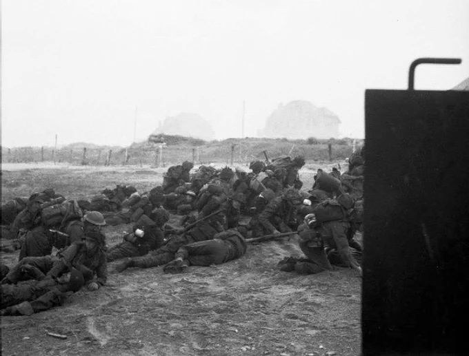 Soldiers lying prone in a training session, wearing helmets and packs, with a blurred vehicle foreground on the right.