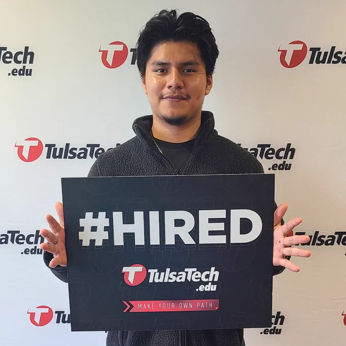 Person holding a #HIRED sign in front of a Tulsa Tech backdrop, celebrating employment success.