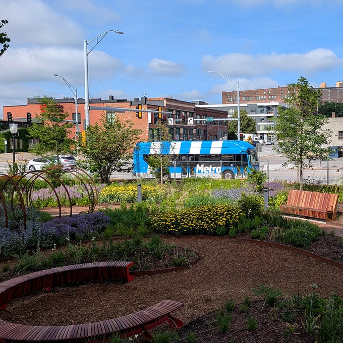 A blue Metro bus drives past the Polina Gardens on 13th Street. The gardens have curved wood benches and many types of flowers and plants.
