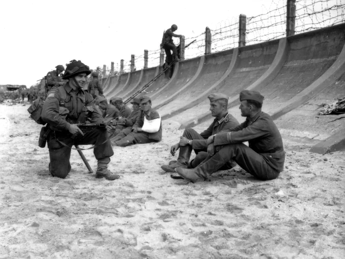 Black and white historical photo depicting a group of military personnel from different nations, sitting and conversing on a sandy beach with barbed wire and structures in the background.