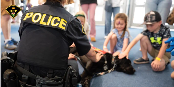 OPP uniformed member with OPP canine at OPP Family and Veterans' Day event. Black Labrador lays down to allow children to pet them.