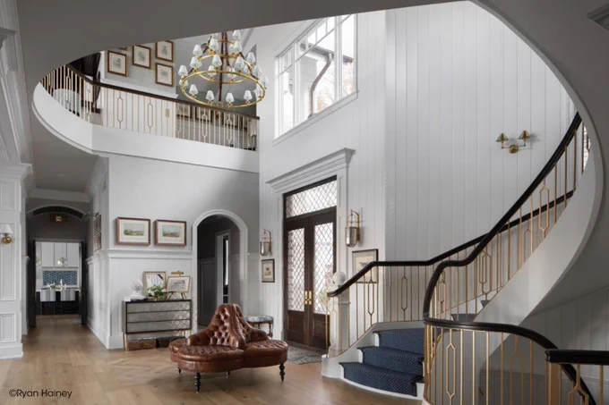 A spacious, elegant interior with a curved staircase, parquet flooring, and a large chandelier. A leather seating area is in the center. Several framed pictures and a decorative mirror adorn the walls. Natural light filters through large windows near the staircase. Copyright Ryan Hainey.