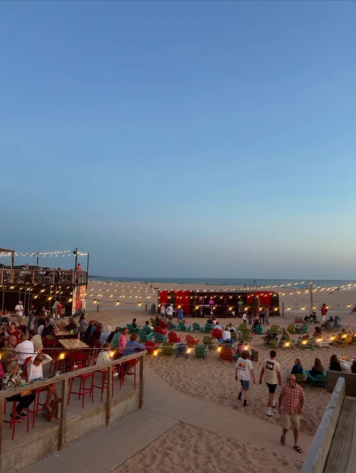 A lively beachside scene at night featuring a gathering of people enjoying a performance. String lights create a warm ambiance. There are colorful chairs arranged in front of a small stage. To the left, a seating area with tables and red bar stools overlooks the scene. The sky is clear with a hint of dusk.