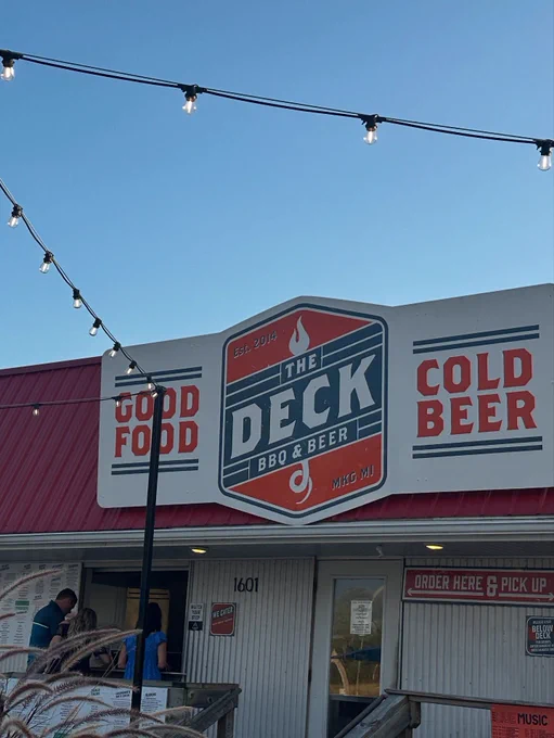The image shows the exterior of "The Deck BBQ & Beer" restaurant, featuring a sign with text including "Good Food" and "Cold Beer." The building has a red roof, and string lights are hanging above. There is a small group of people near the entrance.