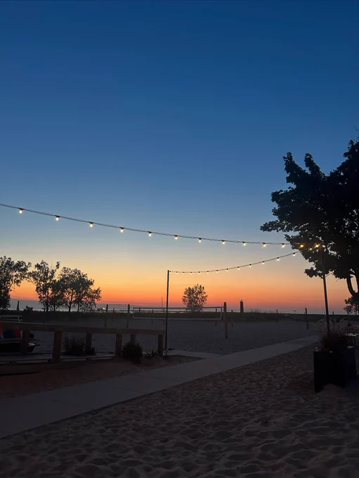 A serene beach scene at sunset with a clear sky transitioning from blue to orange near the horizon. String lights are hanging above, illuminating a sandy path. Trees are silhouetted against the colorful sky, adding to the tranquil atmosphere.