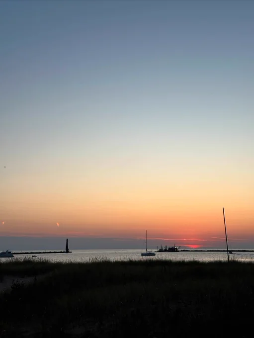 Sunset over Lake Michigan with a silhouette of a lighthouse and boats in the distance. The sky is a gradient of orange, pink, and blue. Grassy foreground.