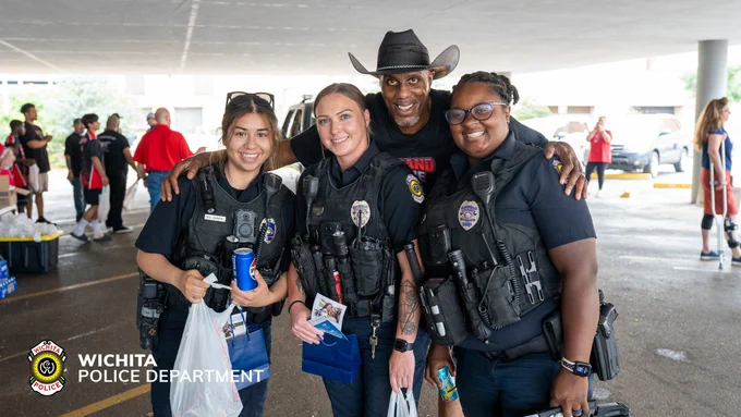 Three Wichita Police Department officers smile and pose with a person wearing a cowboy hat at an event under a large canopy. The officers hold bags and cups, while other people can be seen in the background.