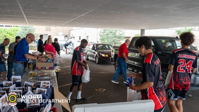 People are gathered under a large canopy for a community event. Several individuals are setting up tables with items. Participants, some wearing sports jerseys, are interacting and distributing goods. Wichita Police Department branding is visible in the corner.