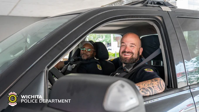 A police vehicle from the Wichita Police Department is parked, with two officers seated inside. The officers are smiling, and the department’s logo is visible on the vehicle.